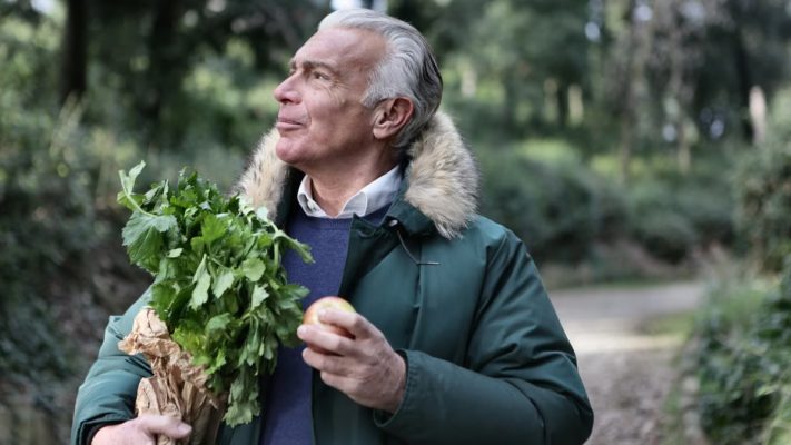 Senior man holding fresh produce and apple, enjoying nature walk.